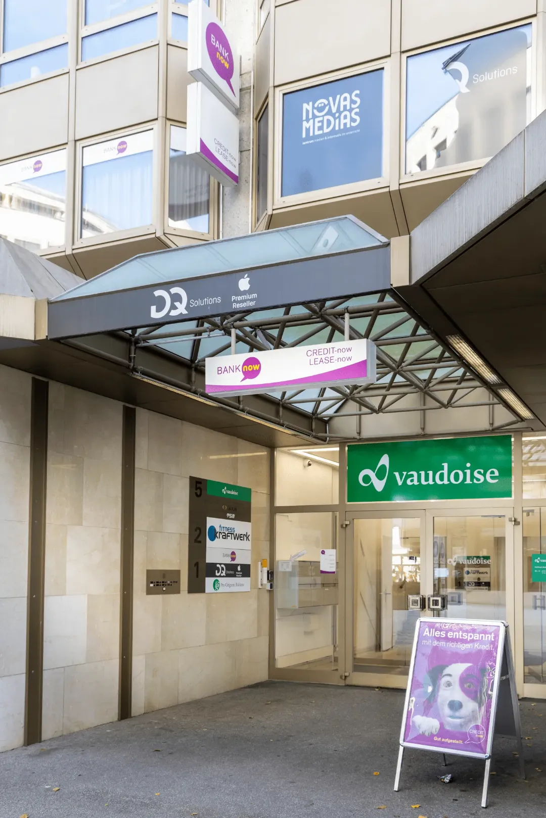 Aussenansicht der BANK-now Filiale Chur mit Eingang unter dem Glasdach und umliegenden Geschäften.