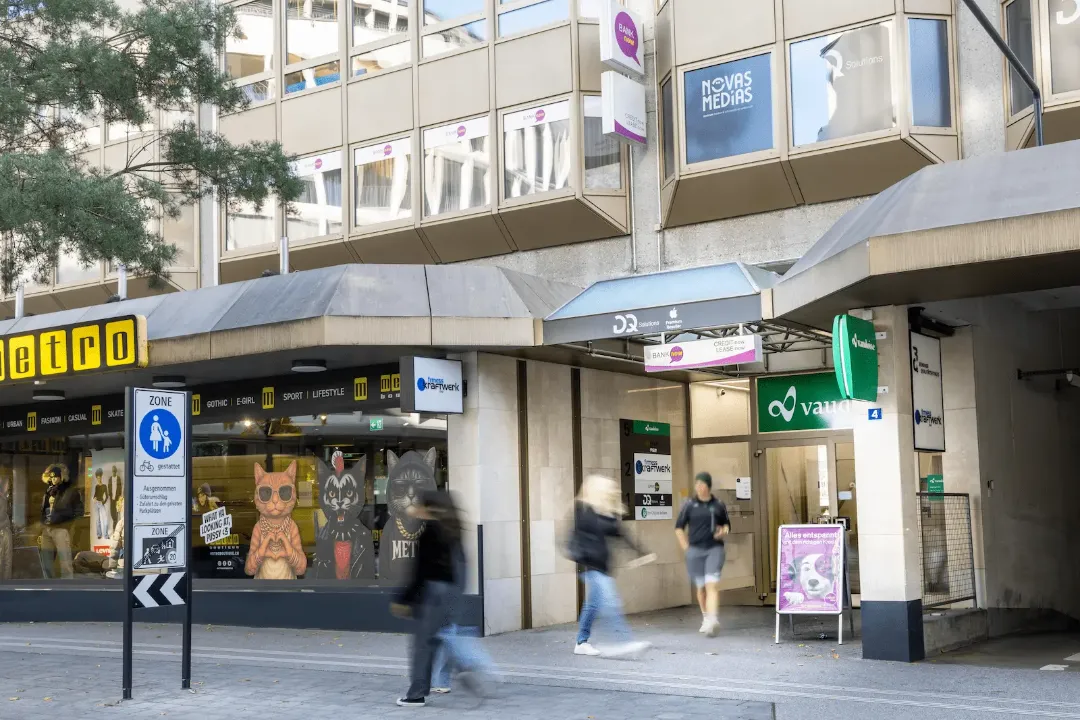 Breiter Blick auf den Eingang der BANK-now Filiale Chur mit Gebäudefassade und umliegender Strasse.
