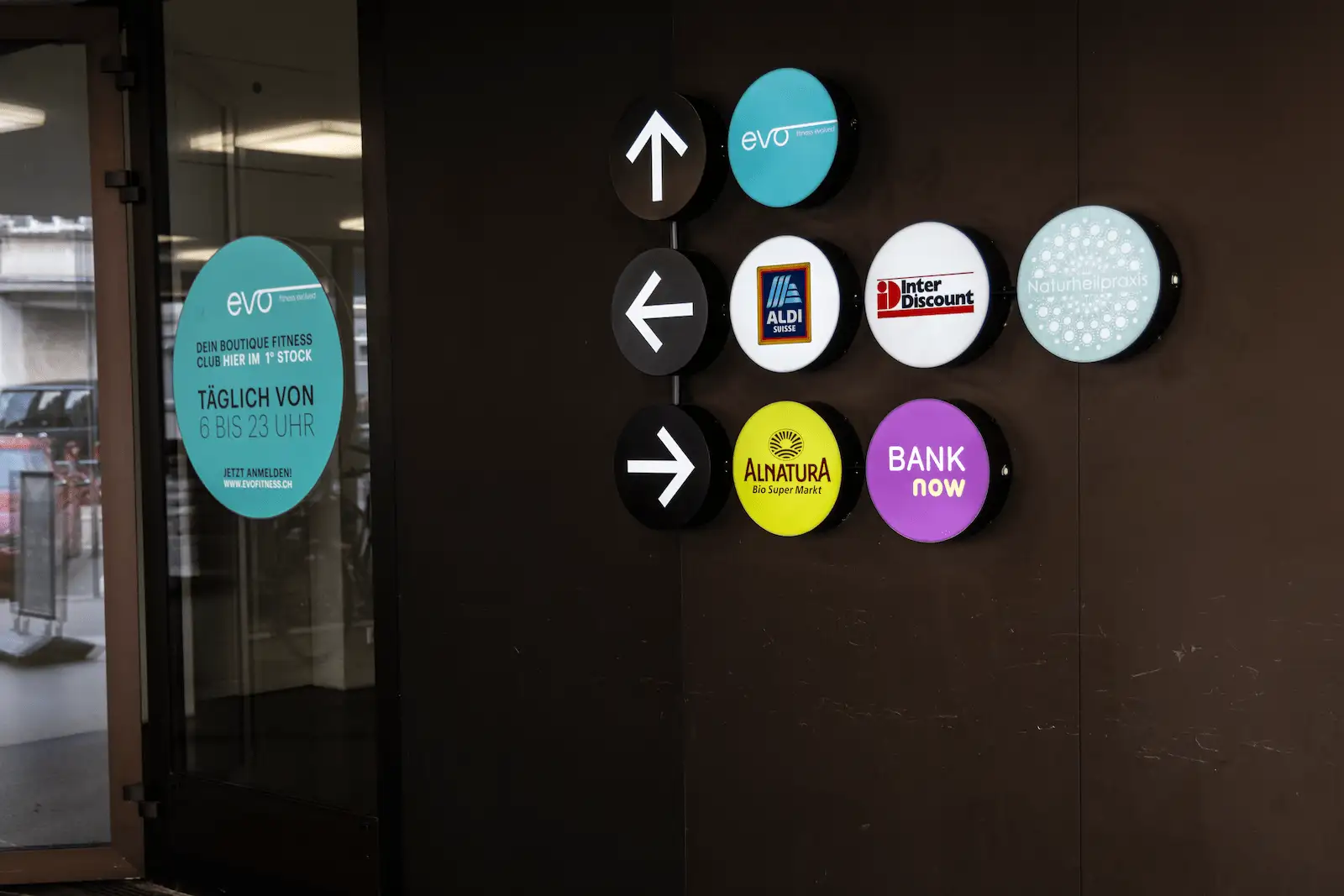 Beschilderung beim Eingang der BANK-now Filiale Zürich-Oerlikon mit Logos und Wegweisern.